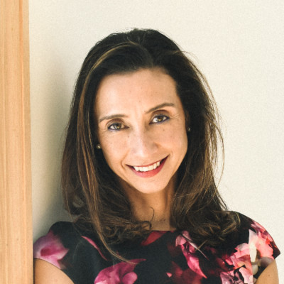 Smiling woman with shoulder-length brown hair wearing a pink floral blouse, standing beside a wooden door.