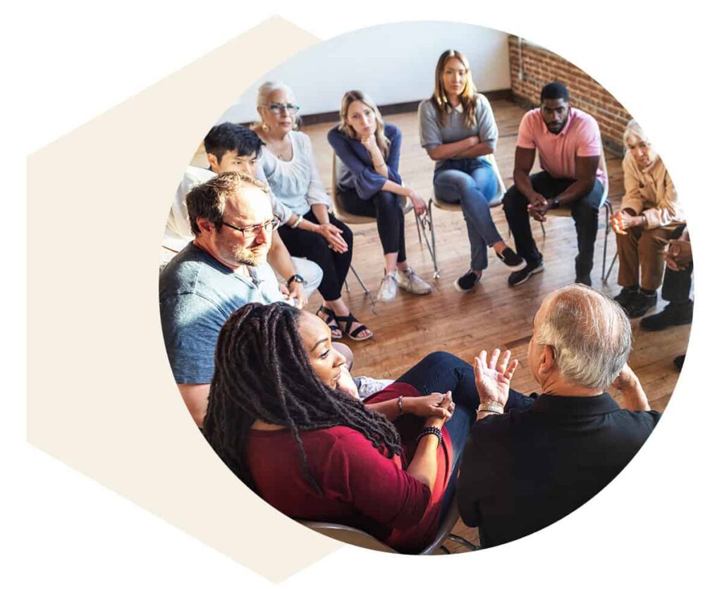 Diverse adults seated in a circle, engaged in a group discussion in a bright room.