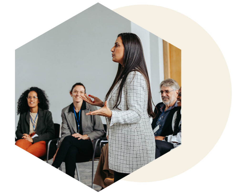Woman in a light tweed blazer speaks and gestures to a seated audience in a conference room.