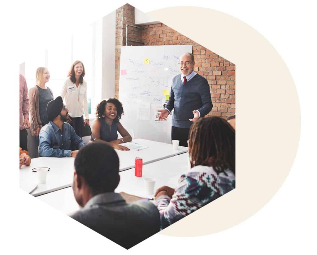 Group of diverse coworkers in a meeting, a man presents at a whiteboard in a brick-walled room.