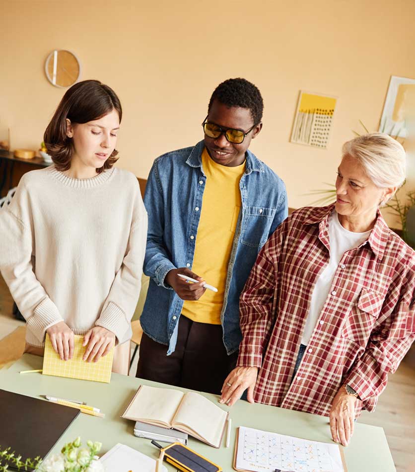 Three people stand around a table with notebooks and a calendar, collaborating on a project.