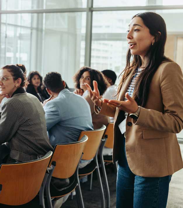 Woman standing and speaking to a group in a conference room with large windows behind them.