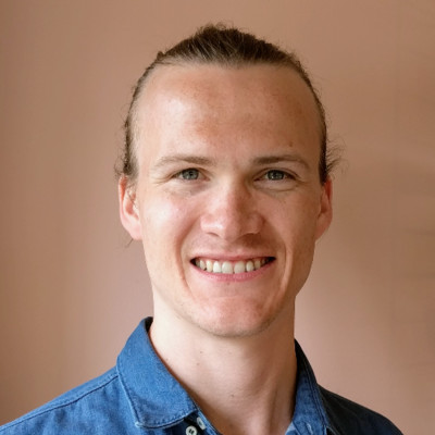 Smiling person with light hair tied back, wearing a blue denim shirt, against a beige background.
