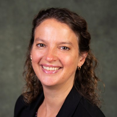 Professional headshot of a smiling woman with dark, wavy hair wearing a black blazer against a neutral backdrop.