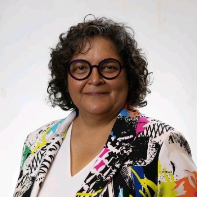 Portrait of a smiling woman with short, curly hair, purple glasses, and a colorful abstract blazer over a white top.