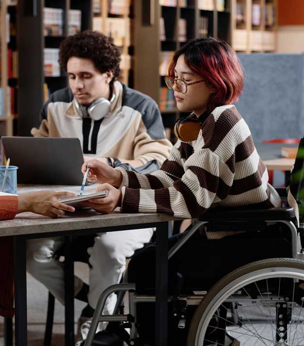 Student in a wheelchair signs a document at a library table while a peer with headphones uses a laptop nearby, studying together.