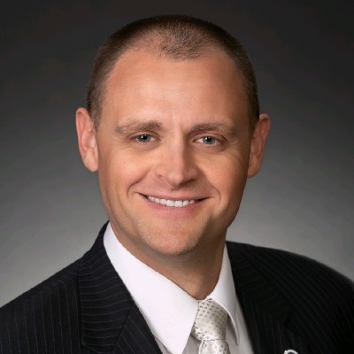 Professional headshot of a smiling man in a dark pinstripe suit and white tie against a gray background.
